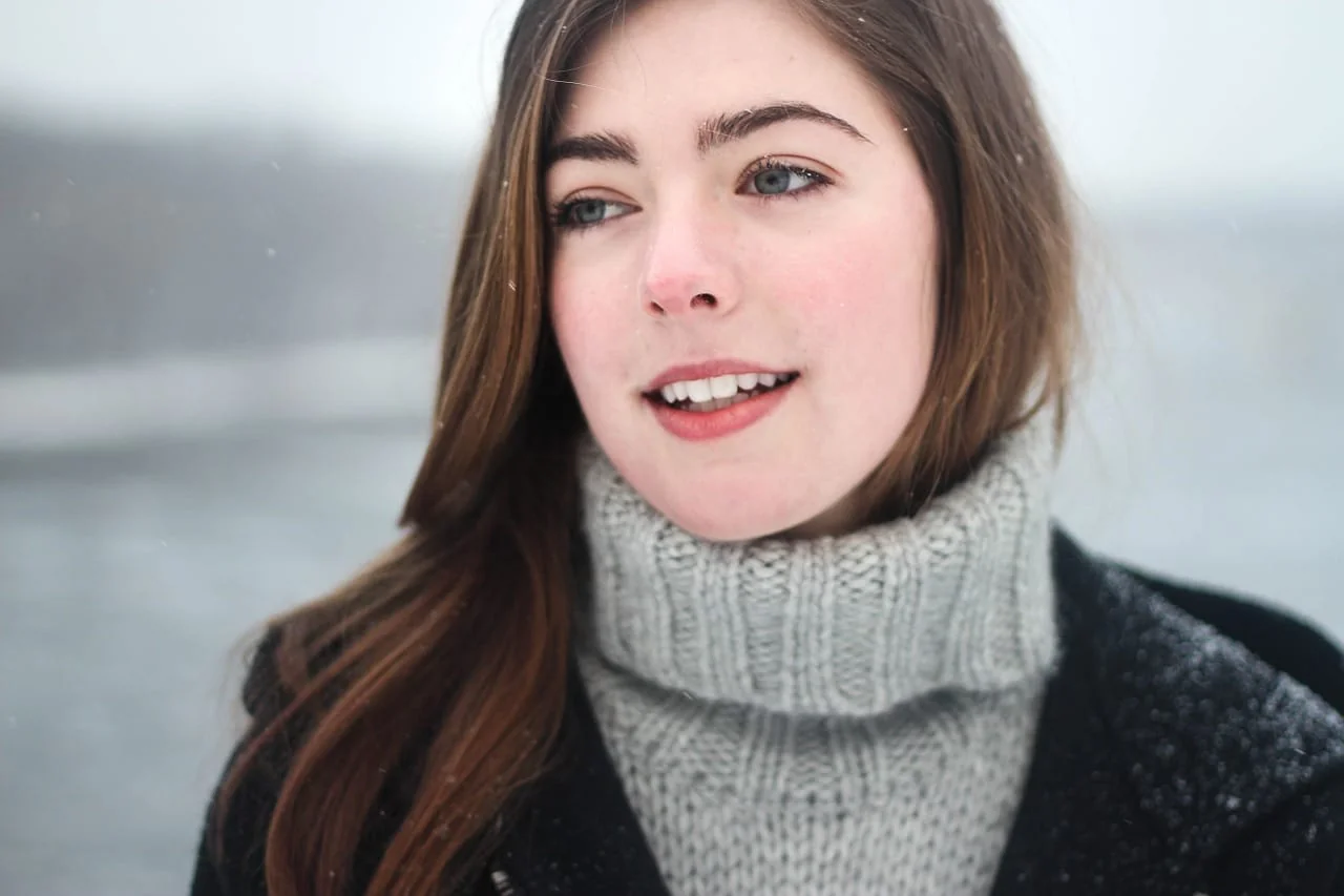 Young woman with long brown hair and a gray turtleneck smiling outdoors in falling snow