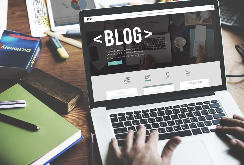 Hands typing on a laptop showing a blog template page with a large "<BLOG>" headline, notebooks and pens on a wooden desk
