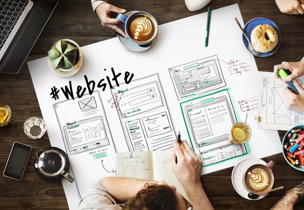 Overhead view of a team sketching website wireframes on a large sheet labeled "#Website", with coffee cups around it