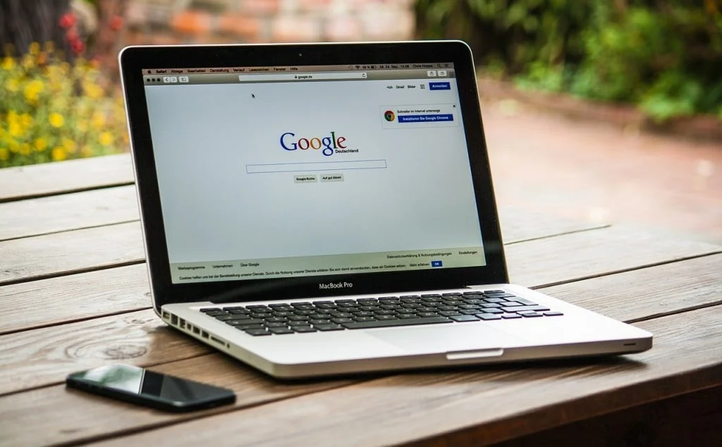 Laptop on an outdoor wooden table showing the Google search homepage, with a phone beside it