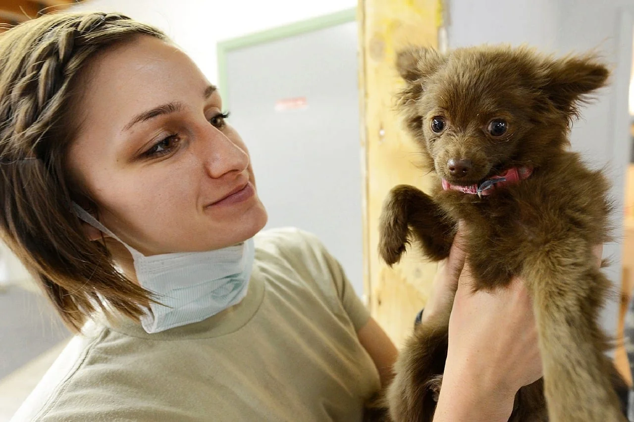 Young woman in a surgical mask pulled down smiling at a small brown fluffy puppy she holds up