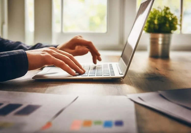 Hands typing on a laptop at a sunlit desk with printed charts in the foreground