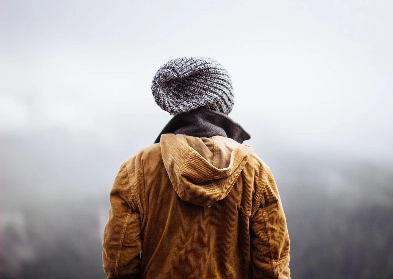 Person seen from behind in a knit beanie and tan corduroy jacket, looking out over a foggy landscape