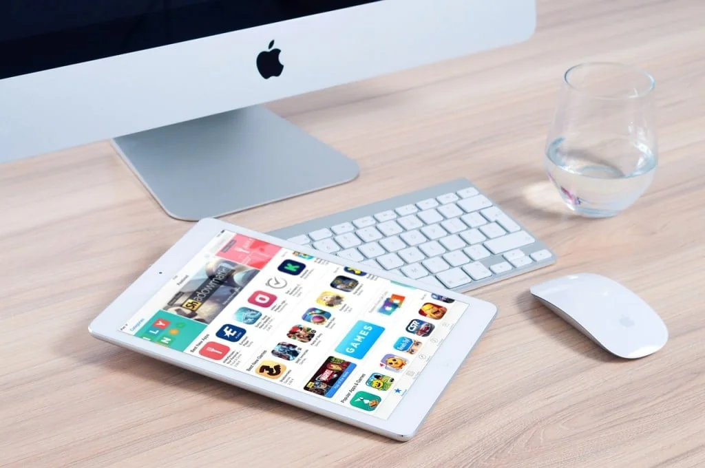 Tablet showing an app store beside a desktop computer, keyboard, mouse and glass of water