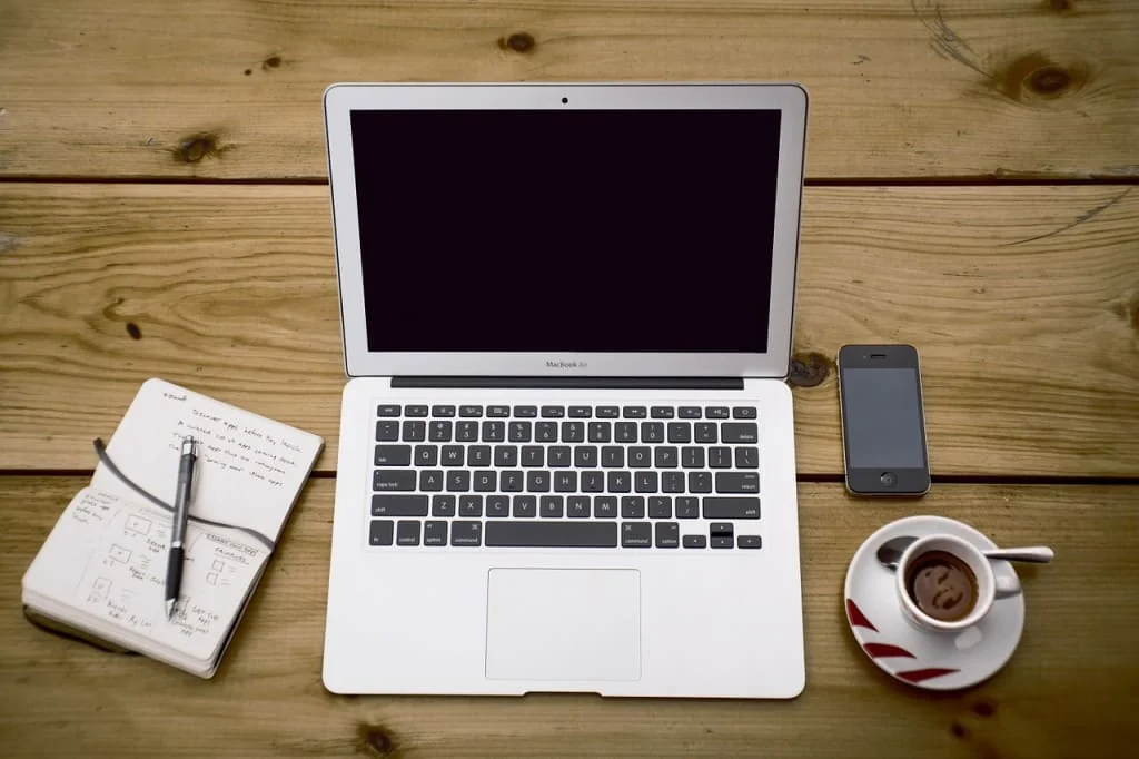 Overhead view of a laptop, smartphone, notebook with pen and espresso cup on a wooden table