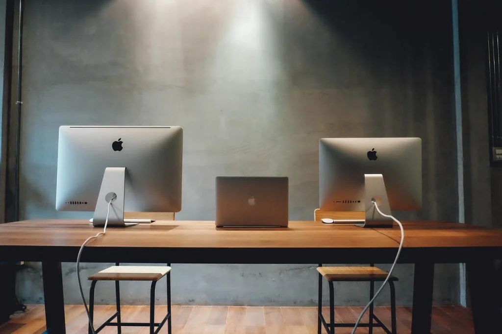 Two desktop computers and a laptop on a long wooden table against a concrete wall, seen from behind
